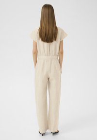Combinaison en lin beige clair avec des manches courtes, taille froncée et design à jambes larges. Présente une finition texturée et décontractée.