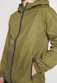 Olivgrön lättviktsjacka med dragkedja framtill, elastiska ärmmuddar och huva. Utrustad med marinblå dragkedjor och en liten logotyp på vänster bröst.