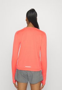 Top sportivo corallo a maniche lunghe con scollo rotondo, texture liscia e dettaglio riflettente sul retro. Abbinato a pantaloni corti grigi.