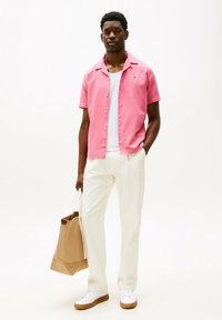 Camicetta rosa a maniche corte con bottoni, canotta bianca, pantaloni crema e borsa a tracolla beige; scarpe bianche con suola marrone.