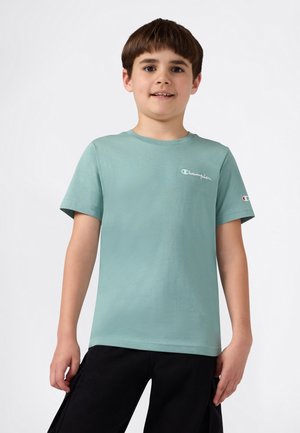 Jongen die glimlacht en een lichtgroene Champion T-shirt en zwarte broek draagt, tegen een effen witte achtergrond.