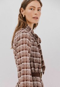 Braun kariertes Kleid mit Kragen und langen Ärmeln, ausgestattet mit einem Gürtel und einem strukturierten Stoff, auf einem Model vor einem neutralen Hintergrund präsentiert.