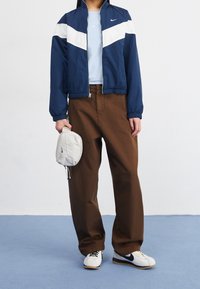 Námořnický modrý větrovka s bílým chevron vzorem, světle modré tričko s kulatým výstřihem, hnědé široké kalhoty a bílé tenisky s černými akcenty.