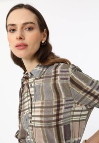 Collared, short-sleeve shirt in plaid design. Features earthy tones of brown, green, and cream with button closure and smooth fabric texture.