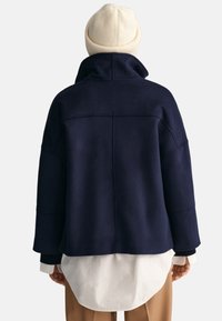 Giacca in misto lana blu navy con colletto alto, spalle dropped e vestibilità rilassata. Presenta una texture liscia e una camicia bianca sovrapposta.