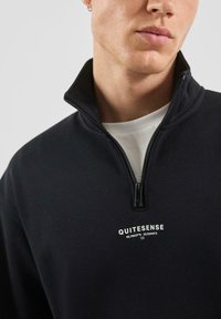 Felpa nera con mezza cerniera e colletto alto, con testo bianco sul petto e una texture liscia e morbida. Dettaglio frontale con cerniera visibile.