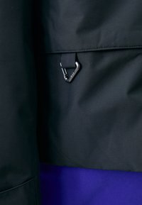 Veste en tissu noir avec une texture mate, dotée d'un clip carabiner noir et d'une doublure violette, mettant l'accent sur un design minimaliste et utilitaire.