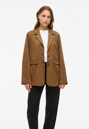 Jeune femme portant un blazer surdimensionné en daim marron, une chemise blanche et un pantalon noir, se tenant devant un fond blanc uni.