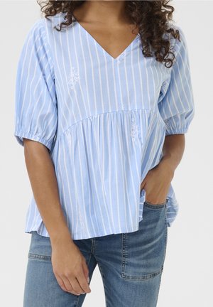 Kobieta w jasnoniebieskiej bluzce w paski z bufiastymi rękawami i niebieskich dżinsach, z ręką częściowo w kieszeni, widoczne kręcone włosy.