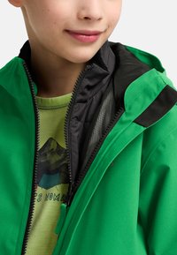 Groene outdoorjas met zwarte accenten, voorzien van een ritssluiting en een meshvoering. Draagt over een lichtgroen graphic t-shirt.