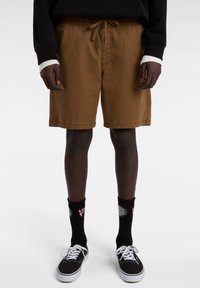 Bruine shorts met een elastische tailleband en een strik. Draagt bij zwarte sokken met een bloemenontwerp en zwarte schoenen met witte veters.