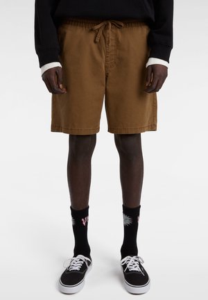 Bruine shorts met een elastische tailleband en een strik. Draagt bij zwarte sokken met een bloemenontwerp en zwarte schoenen met witte veters.