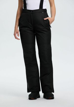 Personne portant un pantalon de neige noir isolé avec des bretelles et des bottes d'hiver noires, se tenant contre un fond uni clair.