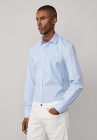Camicia a righe blu chiaro con bottoni, realizzata in cotone, dotata di colletto, maniche lunghe e una vestibilità aderente. Indossata con pantaloni bianchi.