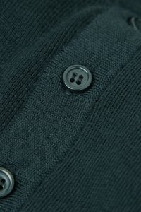 Ciemnozielona dzianina z fakturowanym wzorem. Na brzegu umieszczono dwa czarne guziki z czterema otworami do zapinania.