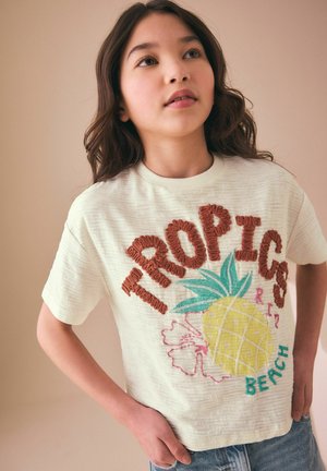 Ung pige med bølget mørkt hår iført en hvid t-shirt med en ananas og teksten "TROPICS BEACH", poserer med hænderne på hofterne.