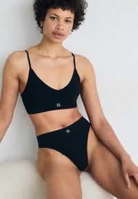 Musta ribattu bralette, jossa on säädettävät hihnat ja logoyksityiskohta, yhdistettynä korkeavyötäröisiin mustiin ribattuihin thong-alushousuihin. Pehmeä rakenne, minimalismi.