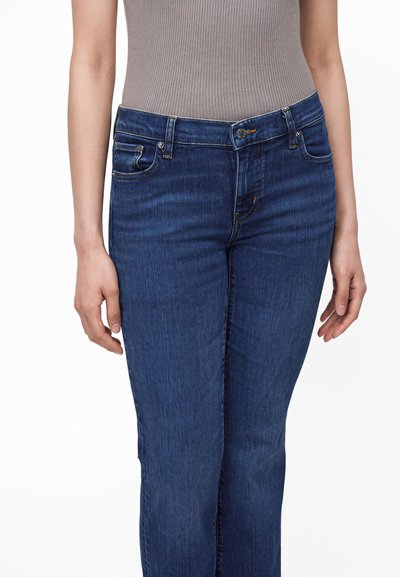 Femme portant un jean en denim bleu et un haut côtelé beige sans manches ajusté, mains détendues le long du corps.