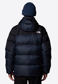 Veste matelassée bleu marine avec manches noires, dotée d'une capuche et de coutures visibles. Comprend un logo de la marque sur le haut du dos à gauche.