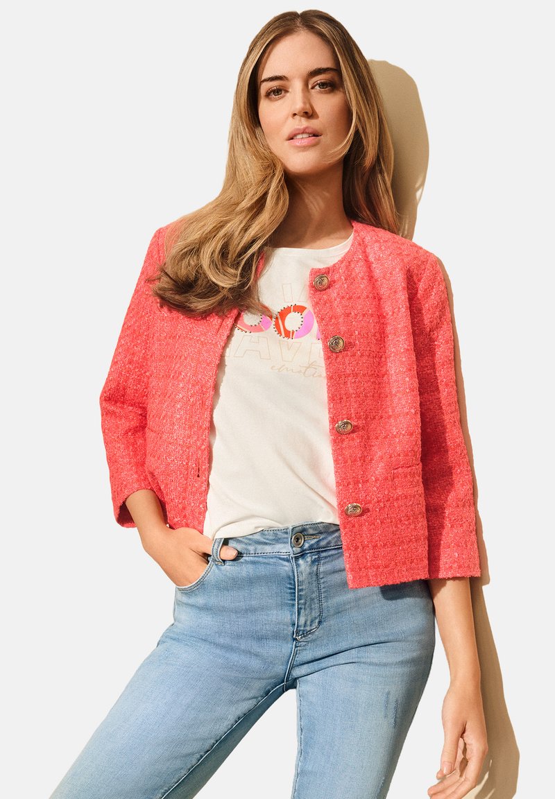 Korall tweed dzseki háromnegyedes ujjal és kerek nyakú kialakítással, arany gombokkal. Fehér grafikai pólóval és világoskék farmernadrággal viselve.