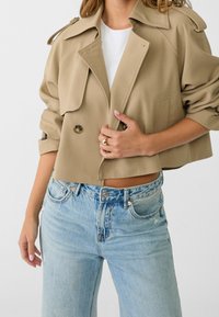 Beżowa, skrócona kurtka z dwurzędowymi guzikami i naramiennikami, zestawiona z jasnoniebieskimi, wysokim stanem jeansami.