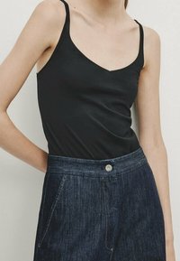 Femme portant un haut noir sans manches en V, rentré dans un pantalon en denim bleu foncé à taille haute avec poches avant et fermeture à bouton.