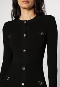 Cardigan noir côtelé à manches longues avec boutons argentés et petites rabats de poche, porté par une personne aux cheveux foncés et au gloss à lèvres neutre.
