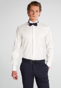 Weißes Hemd mit langen Ärmeln und einer marineblauen Fliege, kombiniert mit dunklen Hosen und einem schwarzen Gürtel. Glatter Stoff mit tailliertem Design.