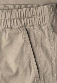 Pas beżowych spodni z elastycznym przeszyciem i widoczną boczną kieszenią z podwójnym szwem w kolorze nici dopasowanym do tkaniny.