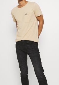 T-shirt beige à col rond avec une poche sur la poitrine gauche, fabriqué en tissu doux, associé à un jean slim fit noir. Design simple et couleur neutre.