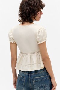 Svetlo béžový top s krátkymi rukávmi, s volánovými rukávmi, priliehavým telom a peplum pásom. Textúrovaná tkanina s jemnými čipkovými detailmi.