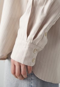 Chemise rayée beige clair à coupe ample, avec poignets boutonnés, texture subtile et tissu doux visible sur la manche.