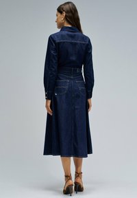 Vestido camisero de denim en azul oscuro con mangas largas, cintura con cinturón y falda acampanada. Presenta dos bolsillos traseros y costuras en contraste.