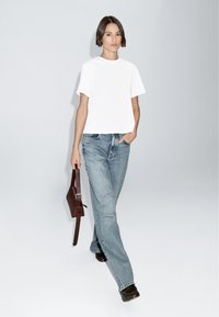 T-shirt bianco a maniche corte abbinato a jeans a vita alta di colore blu chiaro e una borsa in pelle marrone. Il modello è appoggiato a uno sfondo neutro.