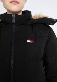 Svart puffjakke med høy krage, glidelås og hette fôret med pels. Har en liten brodert logo i rødt, hvitt og blått.
