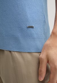 Maglione a maglia blu chiaro con un motivo a punto testurizzato, con una placca logo in argento. Orlo a coste. Abbinato a pantaloni beige.