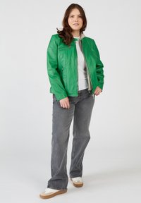 Grön läderjacka med en figursydd design, silverdragkedjor, ståkrage, bärs över en vit skjorta, i kombination med grå jeans och beige sneakers.