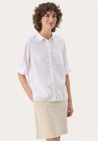Hvid linned skjorte med knapper og rullede ærmer samt en brystlomme, parret med en beige nederdel, der går til knæene.
