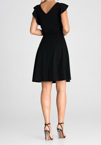 Vestido negro con mangas cortas con volantes y espalda en V, falda acampanada, detalle de cremallera, combinado con sandalias negras de tacón transparente.