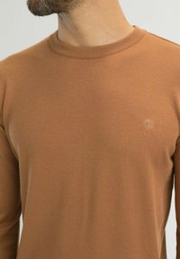 Pull marron à manches longues en tissu tricoté texturé. Il présente un col rond et un petit logo sur la poitrine.