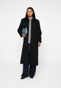 Čierny dlhý kabát z vlnenej zmesi, s ostrým golierom, predným zapínaním na gombíky a oversized rukávmi. Modelka drží malú modrú kabelku.