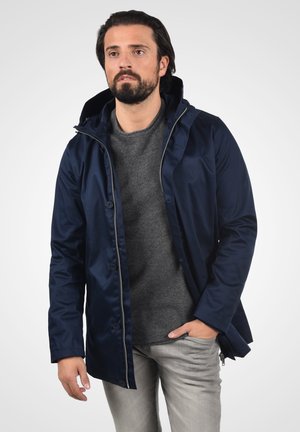 Chaqueta impermeable en color navy con cierre de cremallera y botones, que cuenta con una capucha, textura suave y un brillo sutil, llevada sobre un suéter gris.