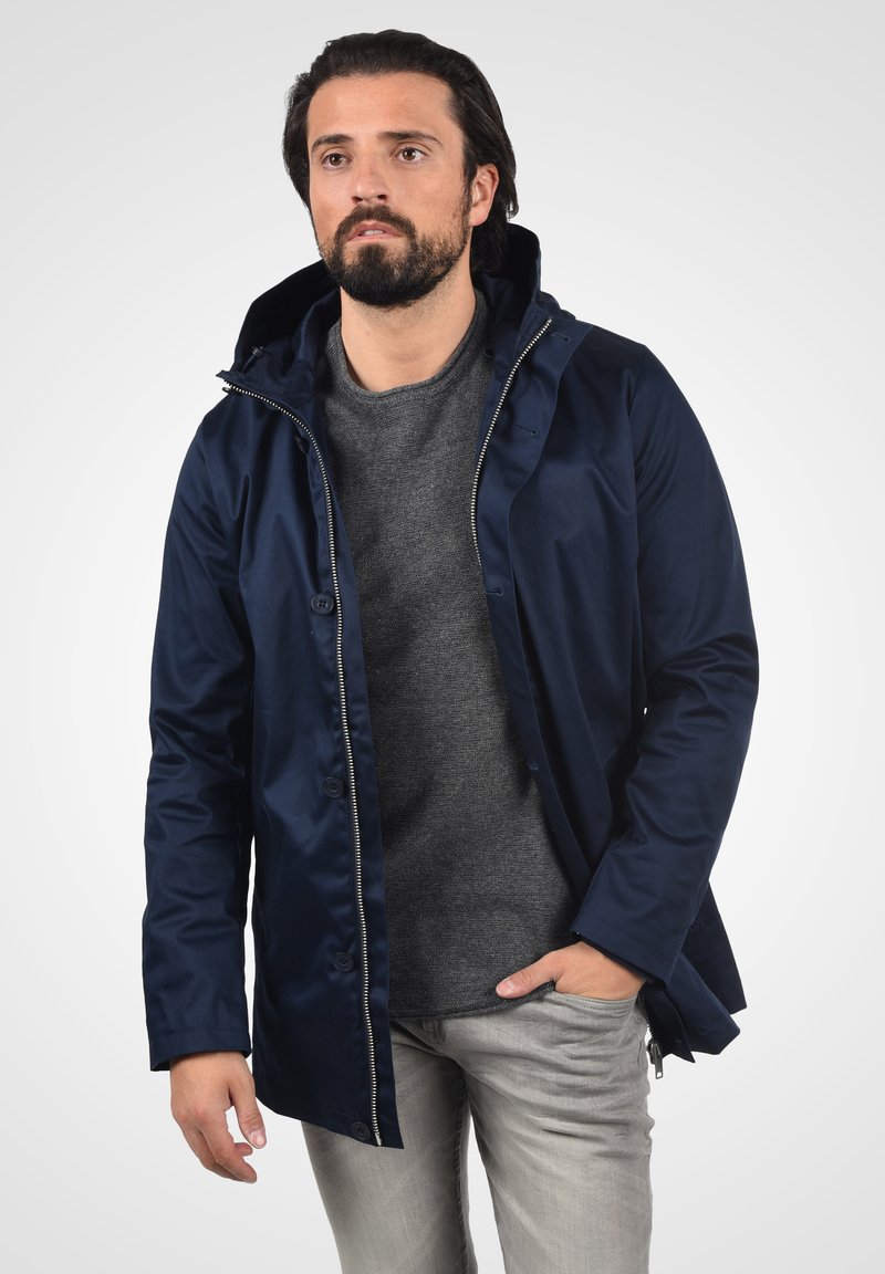Giacca impermeabile navy con chiusura a zip e bottoni, dotata di cappuccio, texture liscia e leggero luccichio, indossata sopra un maglione grigio.