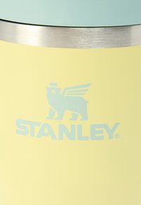 Φωτεινό κίτρινο μονωμένο ποτήρι με καπάκι σε μέντα πράσινο, που διαθέτει ασημί ταινία και μπλε λογότυπο αρκούδας με φτερά και το όνομα της επωνυμίας "STANLEY".