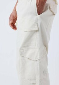 I pantaloni cargo beige presentano diverse tasche laterali, una vestibilità comoda e un dettaglio del logo ricamato vicino alla tasca. Realizzati in tessuto resistente.