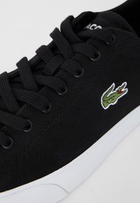 Sneaker nera in tela con lacci, suola bianca e piccolo logo di coccodrillo ricamato verde sul lato. Tessuto testurizzato e design elegante.