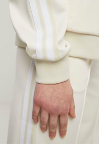Kremfarget hettegenser med teksturerte mansjetter og hvite striper langs ermene. Hånden hviler på matchende bukser med sidelapper.