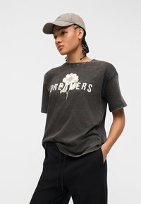 Γυναίκα που φοράει γκρι μπλουζάκι με τη λέξη "DREAMERS" και λευκή εκτύπωση λουλουδιού, μαύρο παντελόνι και ανοιχτό γκρι καπέλο baseball, με τα χέρια στις τσέπες.