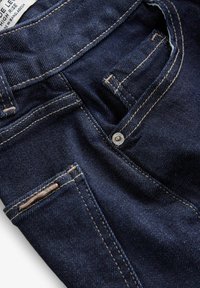 Gros plan de la zone de poche avant d'un jean en denim bleu foncé avec couture beige et petit détail de bouton à rivet métallique.