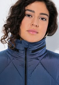 Kvinna med lockigt mörkt hår som bär guldörhänge och en blå puffjacka med hög krage, allt mot en vit bakgrund.